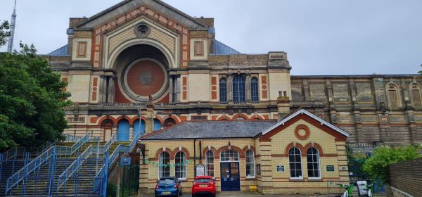 In the foreground is Alexandra Palace station, a one-storey cream brick building with burnt orange arches. Immediately behind it is Alexandra Palace, a much bigger building in the same style. To the left of the station building is some steps up to Alexandra Palace, with blue railings and fencing.