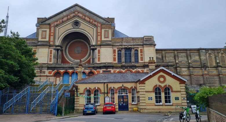 In the foreground is Alexandra Palace station, a one-storey cream brick building with burnt orange arches. Immediately behind it is Alexandra Palace, a much bigger building in the same style. To the left of the station building is some steps up to Alexandra Palace, with blue railings and fencing.