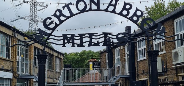 Iron arch, on which is an engraved sign stating Merton Abbey Mills. Behind this sign are two storey buildings to the left and right. An electricity pylon can be seen in the distance.