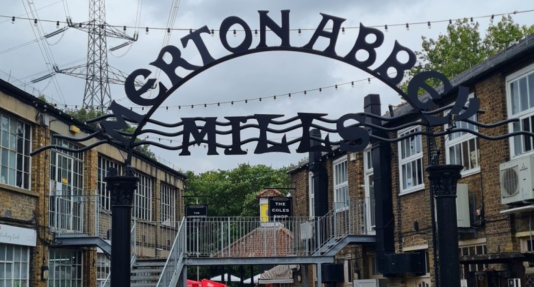 Iron arch, on which is an engraved sign stating Merton Abbey Mills. Behind this sign are two storey buildings to the left and right. An electricity pylon can be seen in the distance.
