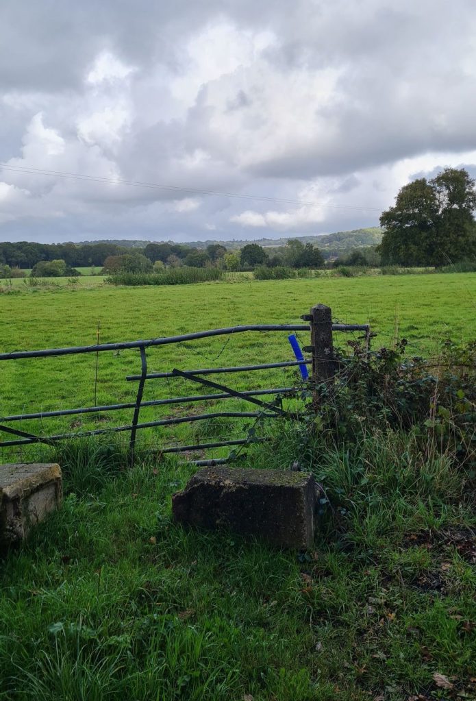 Looking out from the side of the path. In the foreground is a partially knocked over gate. In the background is a large green field, beneath a grey and cloudy sky.