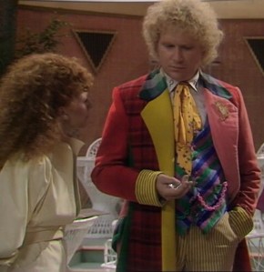 The Doctor and Mel in a scene in the 1986 TV story Terror of the Vervoids. Mel is turned to one side facing the Doctor, with curly red hair and a cream robe, and the Doctor is looking puzzled, wearing a rainbow coat and a yellow cravat.